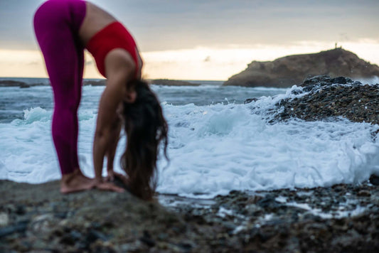 A Yogini on a rocky beach does a standing forward bend with a crashing ocean in background.